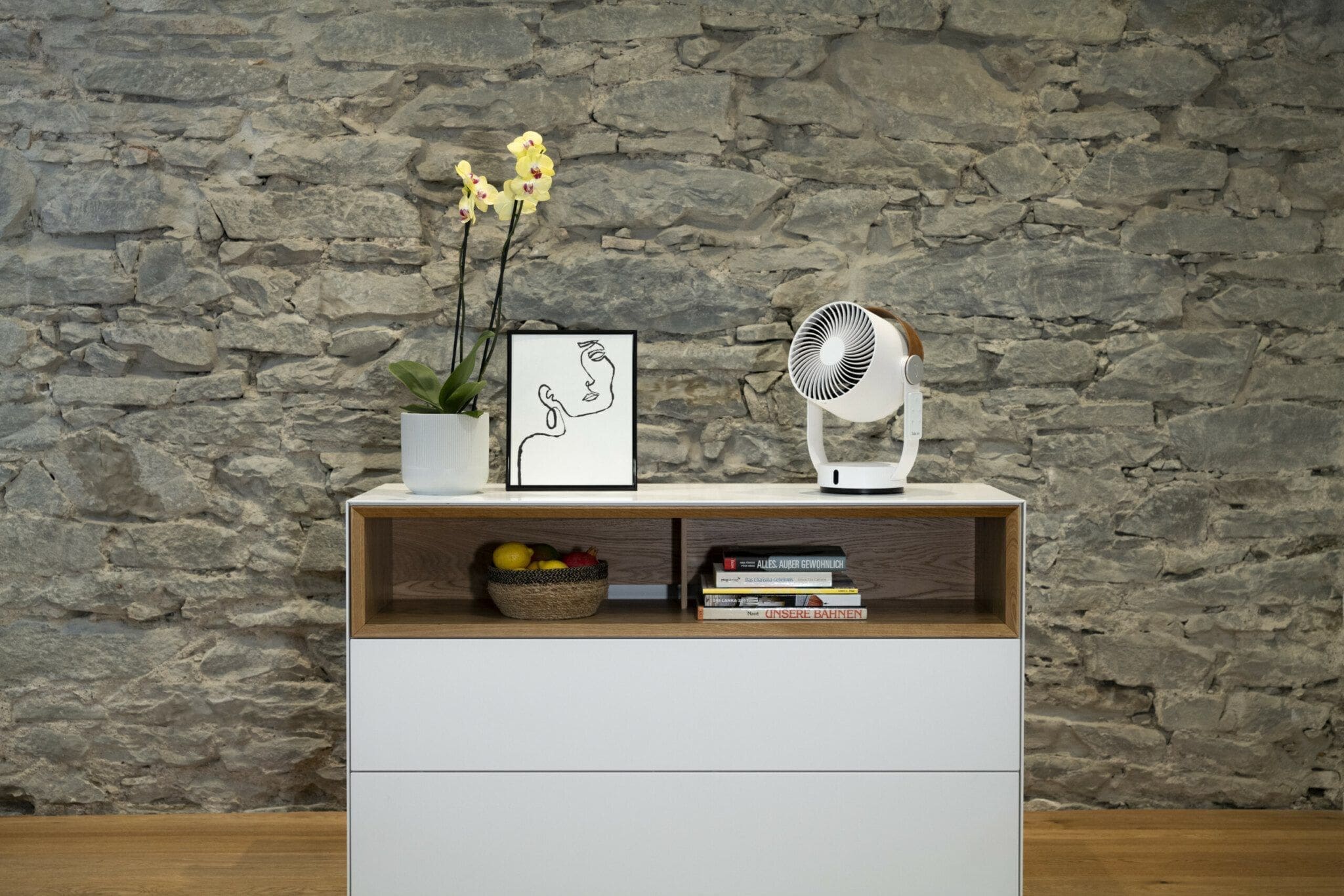 Weißer SF Leo Ventilator auf Kommode. Deko mit Orchidee, Bild, Obstkorb und Bücher. Steinwand im Hintergrund.