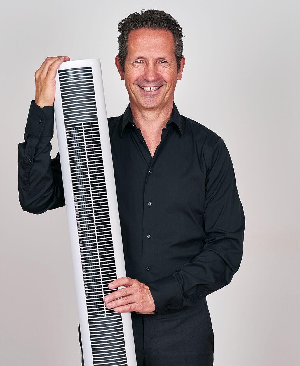 Peter White Martin präsentiert einen Turmventilator. Der Ventilator ist weiß und schlank, der Mann lächelt. Kühles Design für heiße Tage.