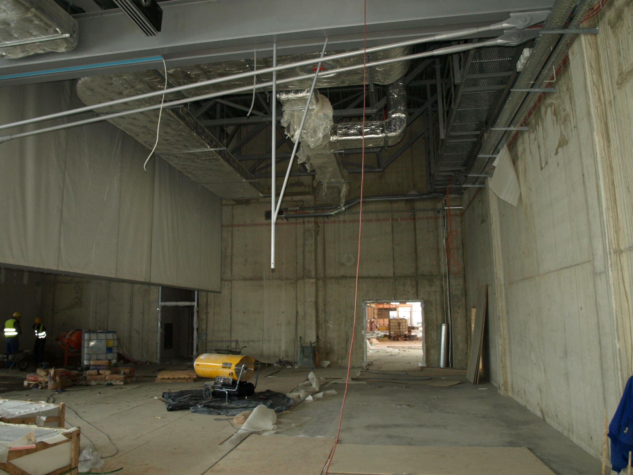 Flughafen Baustelle Posen: Innenansicht einer Halle im Rohbau. Betonwände, Deckeninstallationen und Baumaschinen sichtbar. Bauarbeiter im Hintergrund.