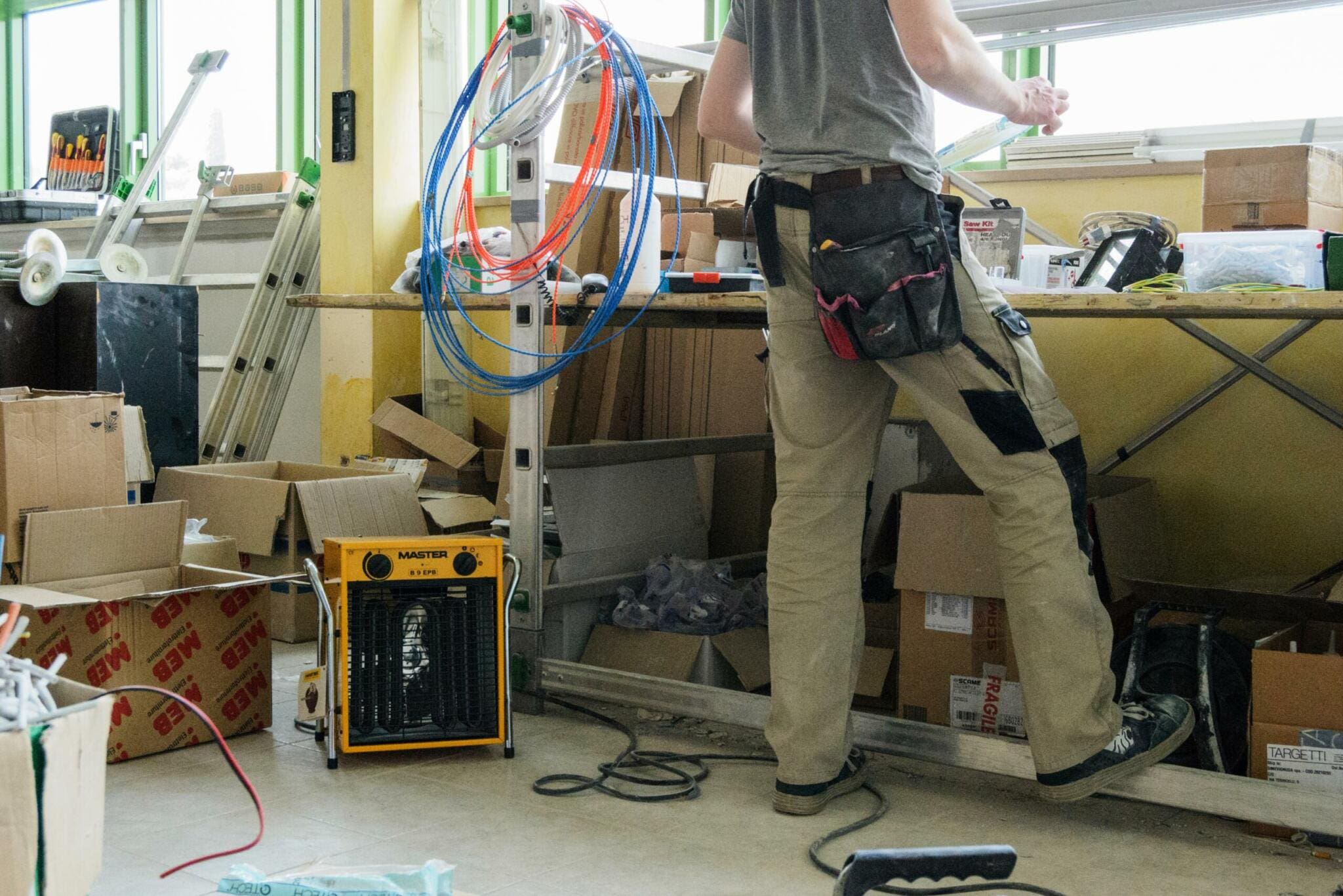 Handwerker im Master Workshop. Werkstatt mit Werkzeug, Kabeln, Leiter und Kartons. Master B-9 Heizlüfter im Vordergrund.