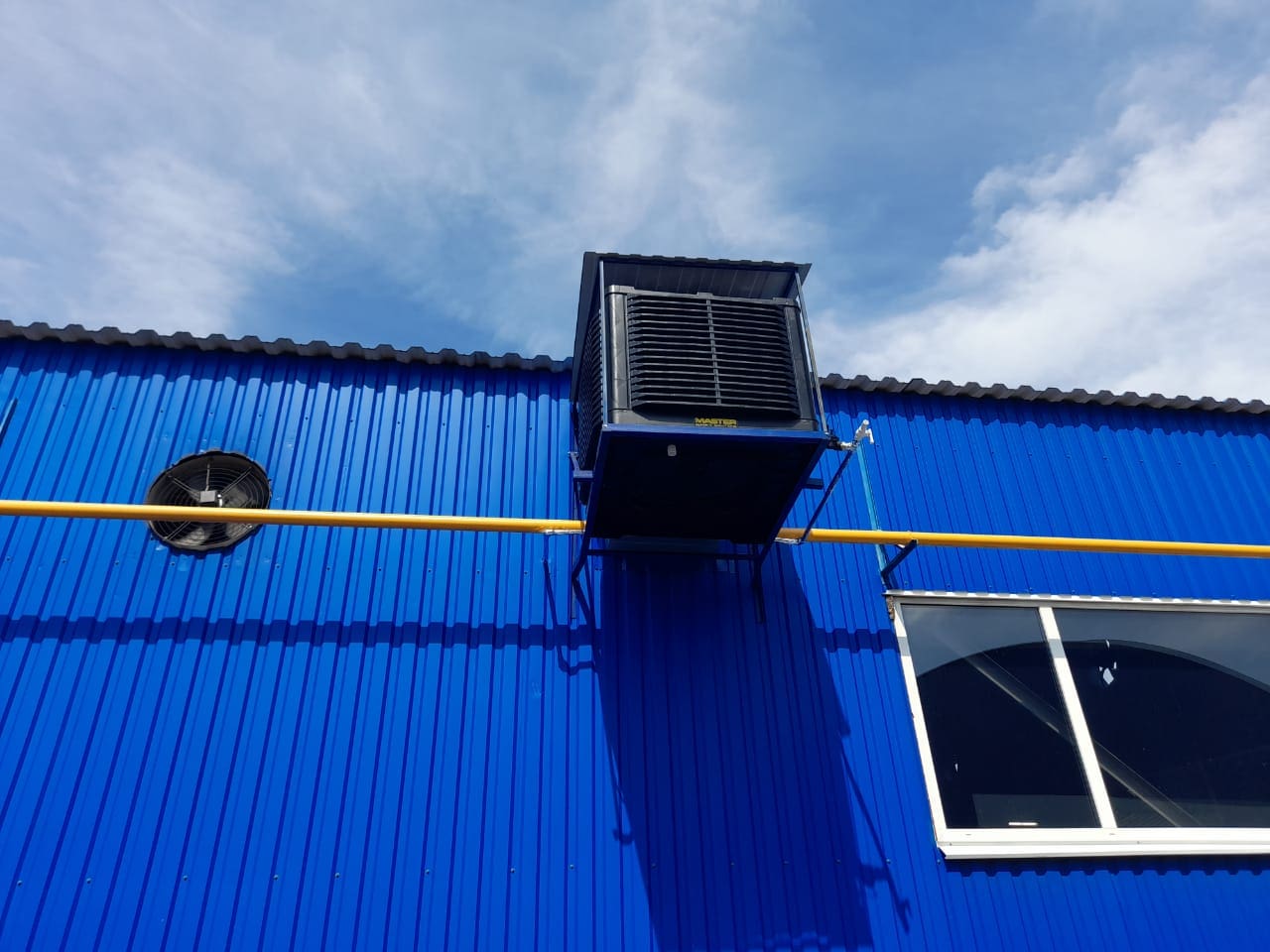 Master BCM Seitenwand-Installation: Kühler an blauer Wand mit Fenster und gelbem Rohr. Ventilator sichtbar. Außenaufnahme.