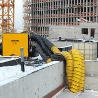 Winterbaustelle: Gelber Master BV 691 Heizlüfter auf einem Gebäude, Bauarbeiten im Hintergrund. Schnee liegt.