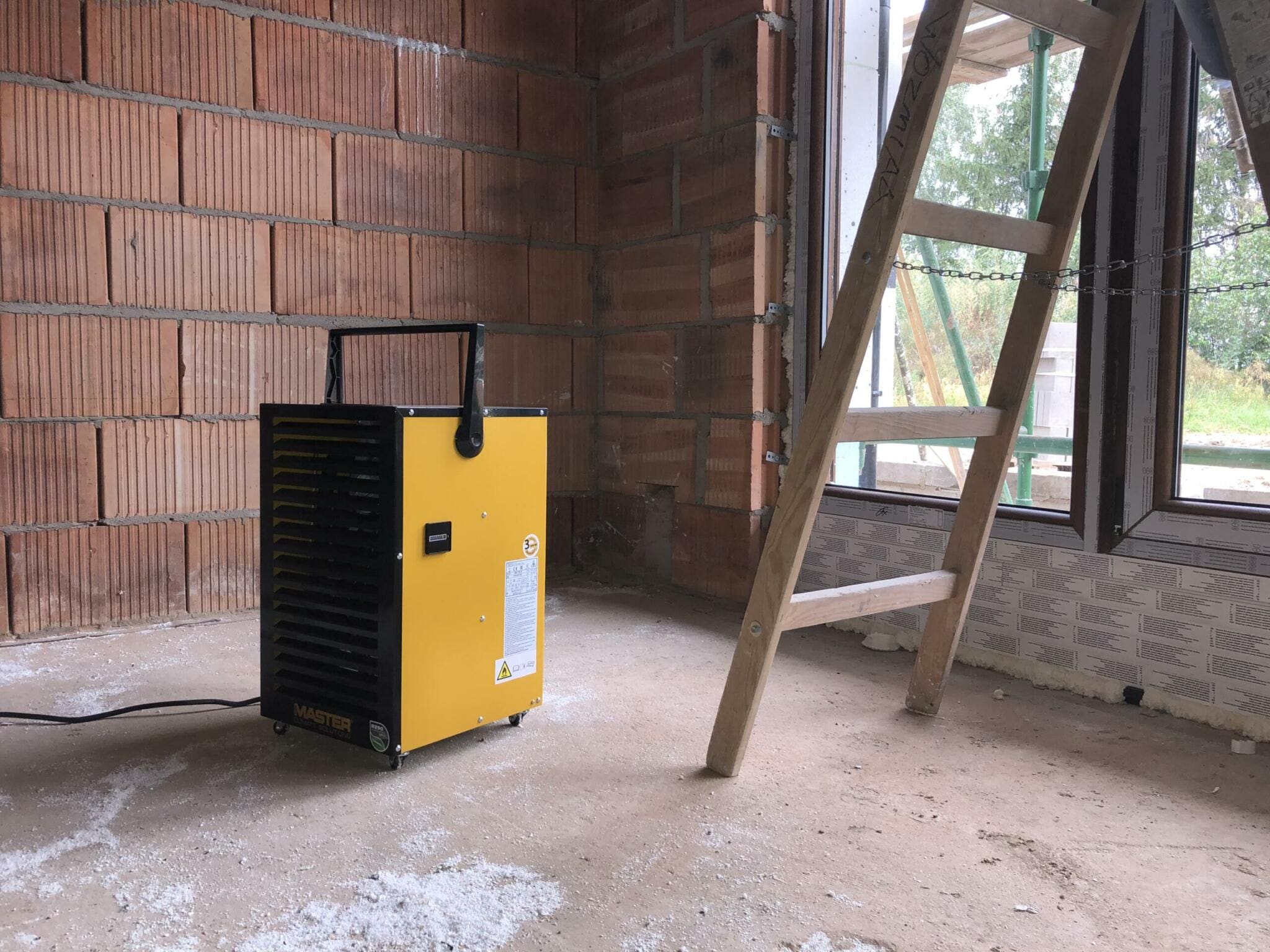 Baustelle: Gelber Master DH 732 Bautrockner neben Leiter vor Ziegelwand und Fenster. Bauarbeiten im Innenraum.