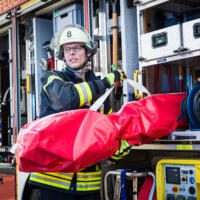 Feuerwehrmann mit roter Tragetasche Typ 180. Einsatzbereit hält er die Tasche, während er im Hintergrund des Einsatzwagens steht.